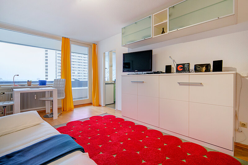 Zu sehen ist ein Einzelbett links, gelbe Vorhänge, ein roter Teppich und ein weißes Sideboard mit Flachbildfernseher.